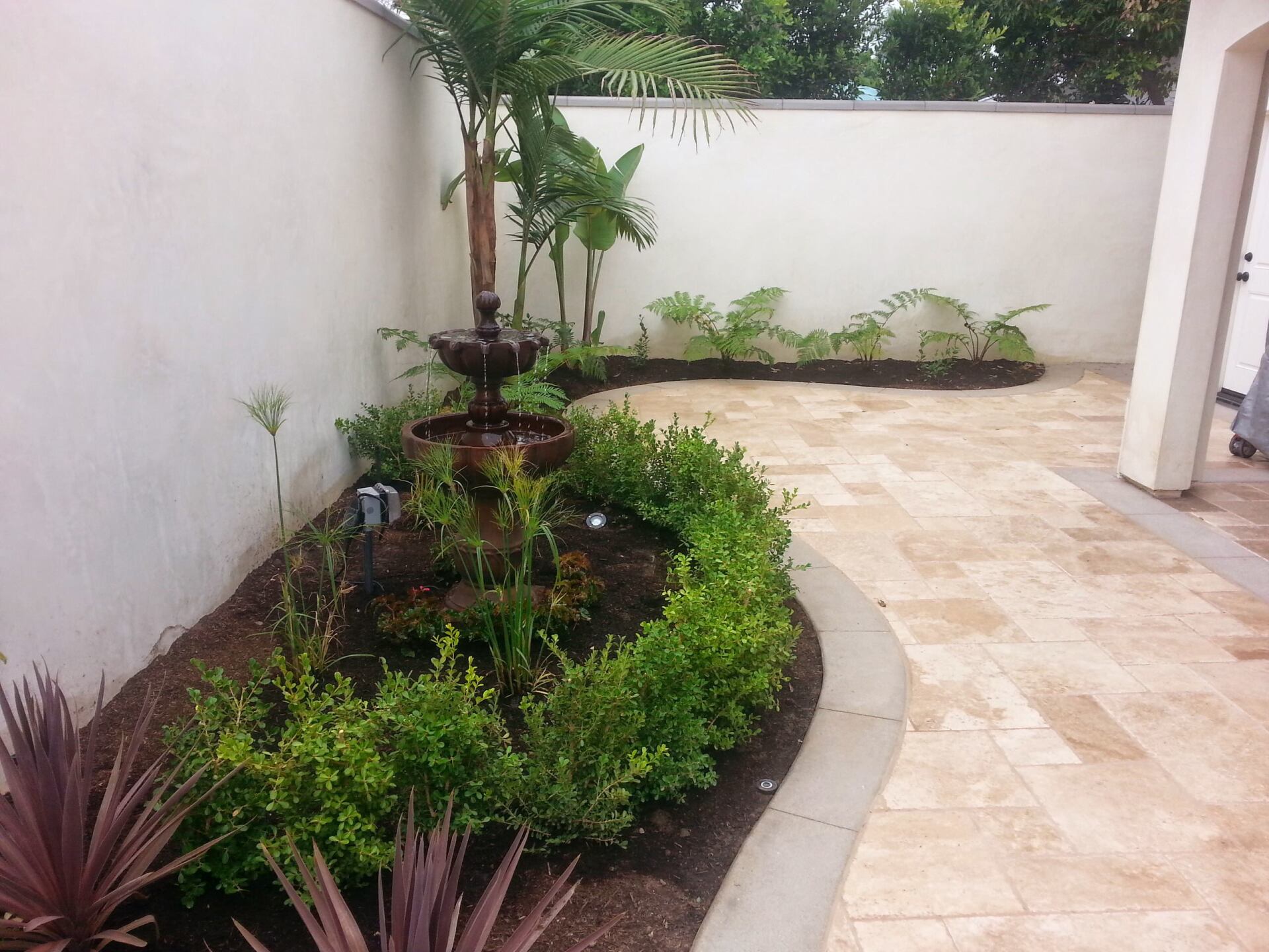 A small courtyard garden with a fountain, palm tree, and neatly trimmed hedges bordering a stone patio.
