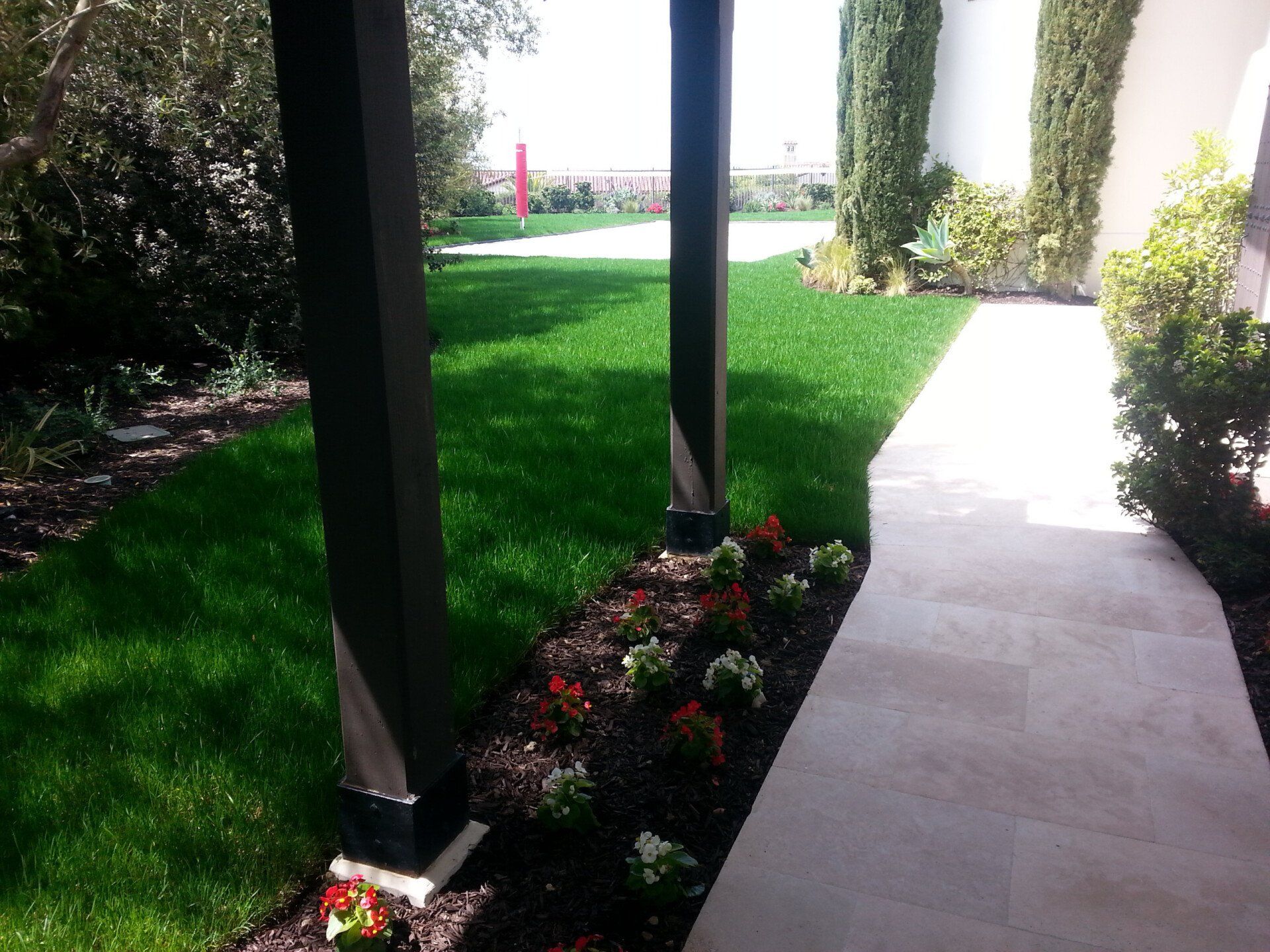 A manicured lawn with flowerbeds, a tiled walkway, and tall columns under a bright sky.