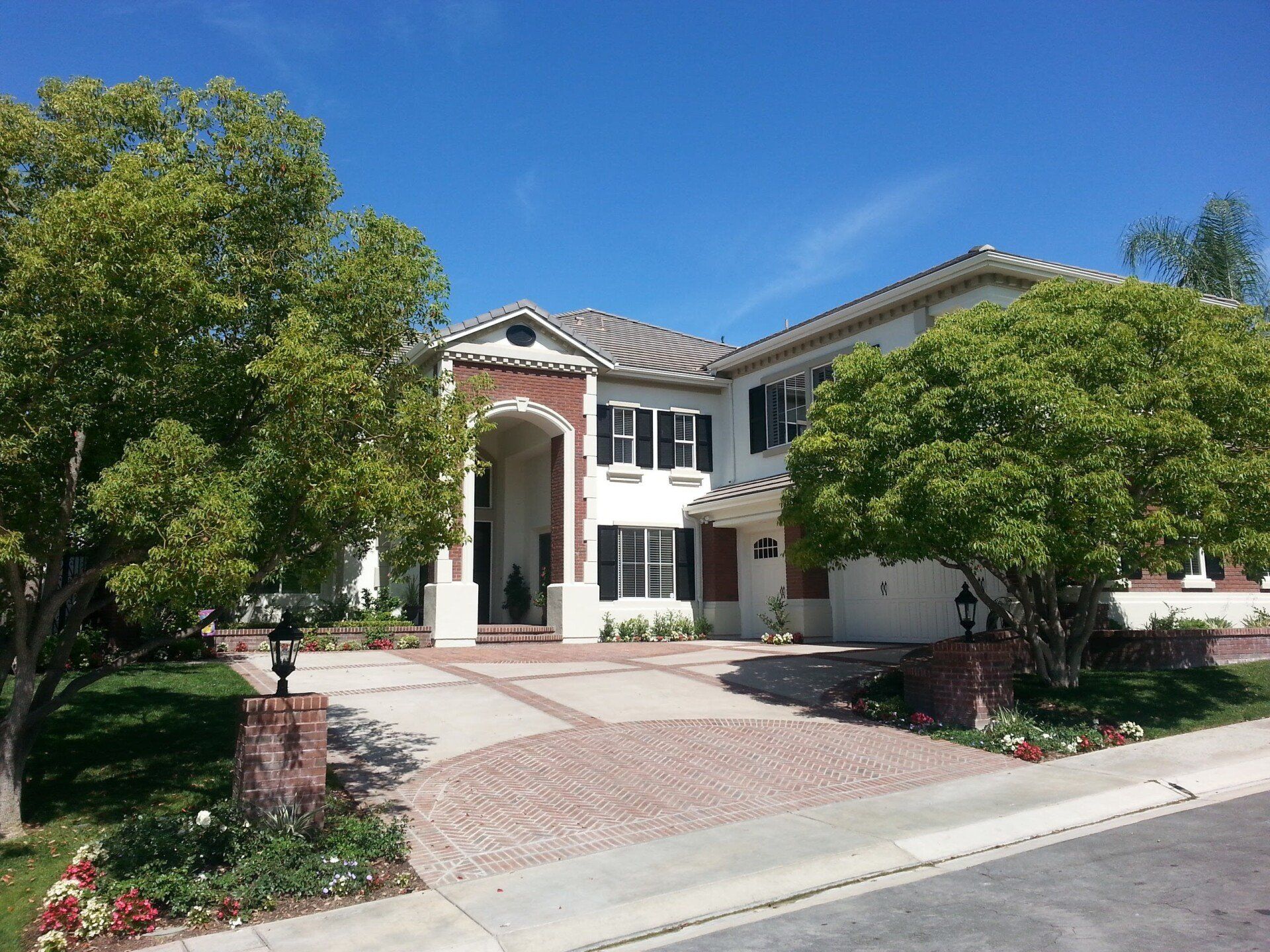 Large white house with brick accents, a patterned driveway, and lush green trees under a blue sky.