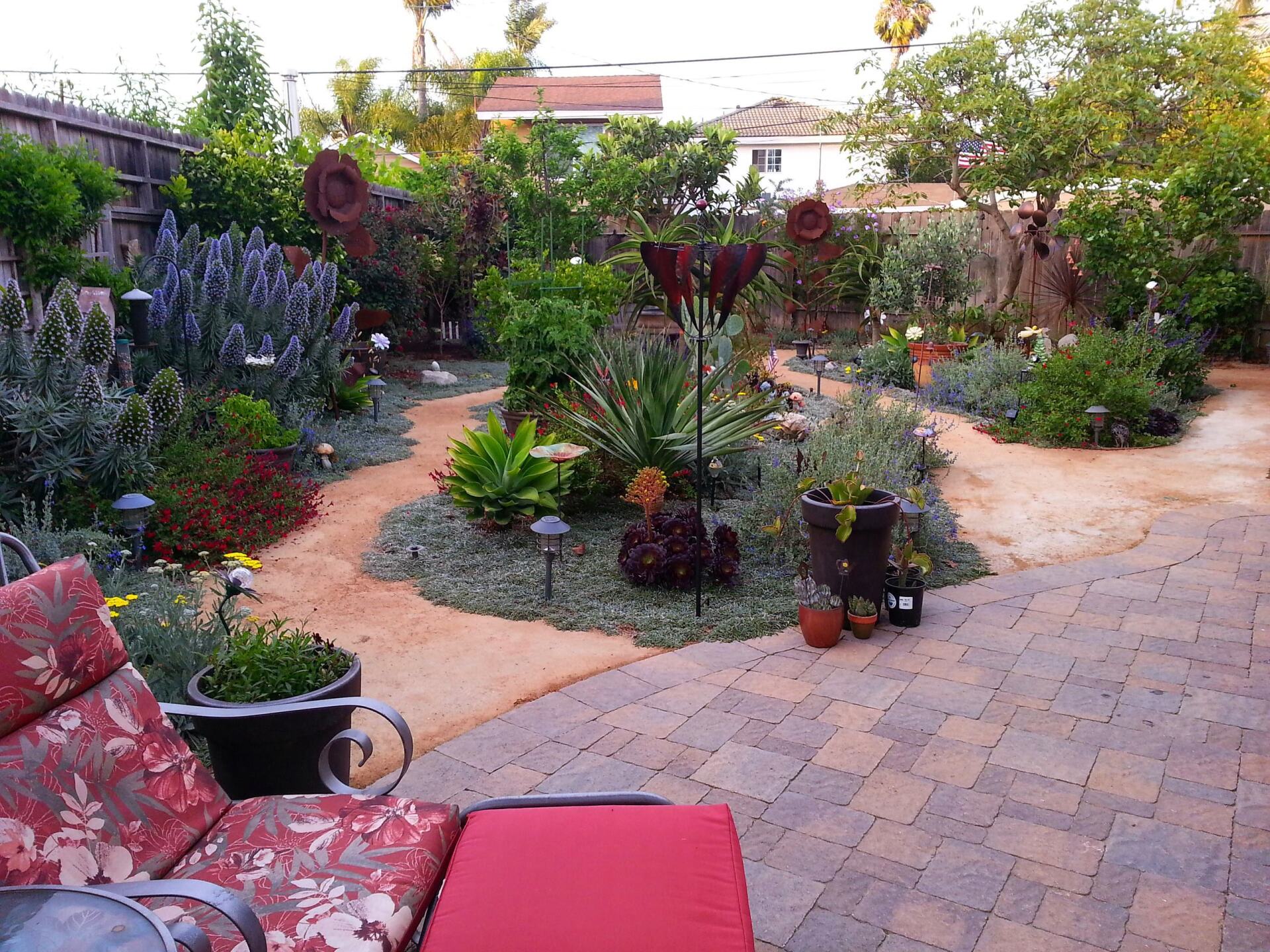 A lush garden with a brick patio and winding pathways, featuring various plants and decorative metal art.