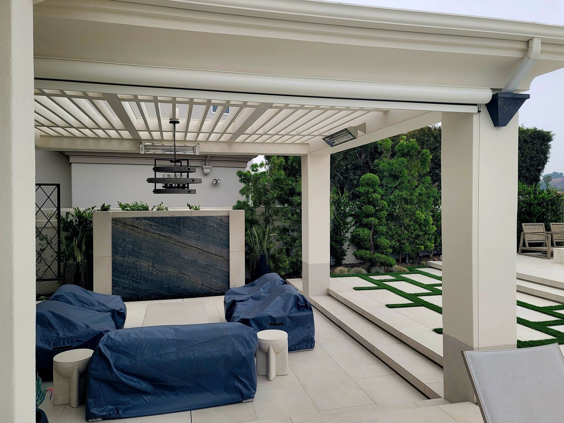 Patio with pergola, covered furniture, water feature, green lawn, and trees.