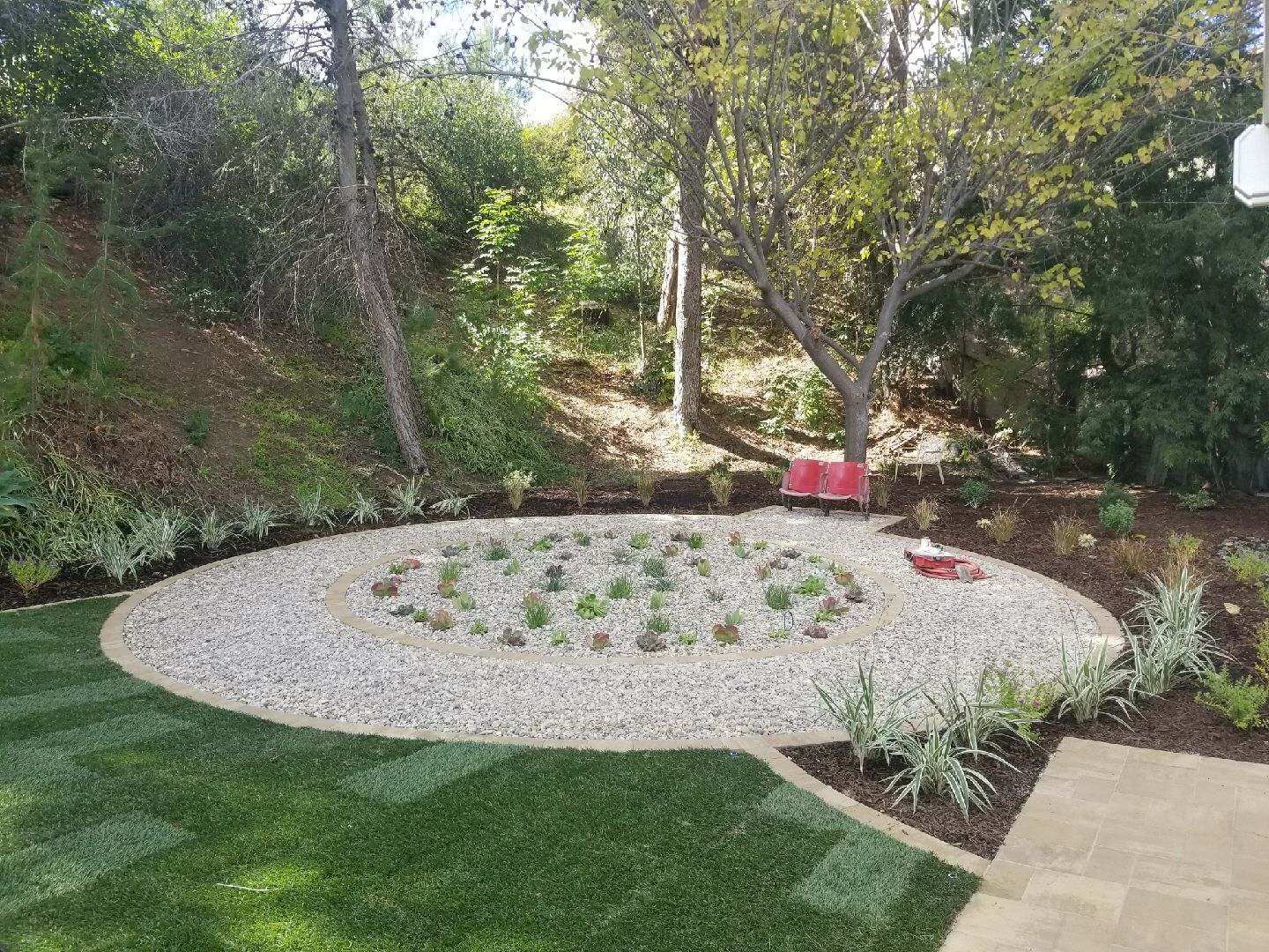 Circular gravel bed with plants, surrounded by lawn and trees, in a backyard setting.