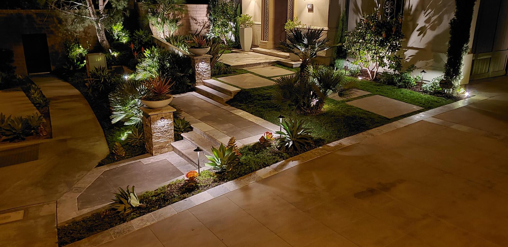 Night view of a house entrance with well-lit landscaping, including plants, pathways, and decorative lights.