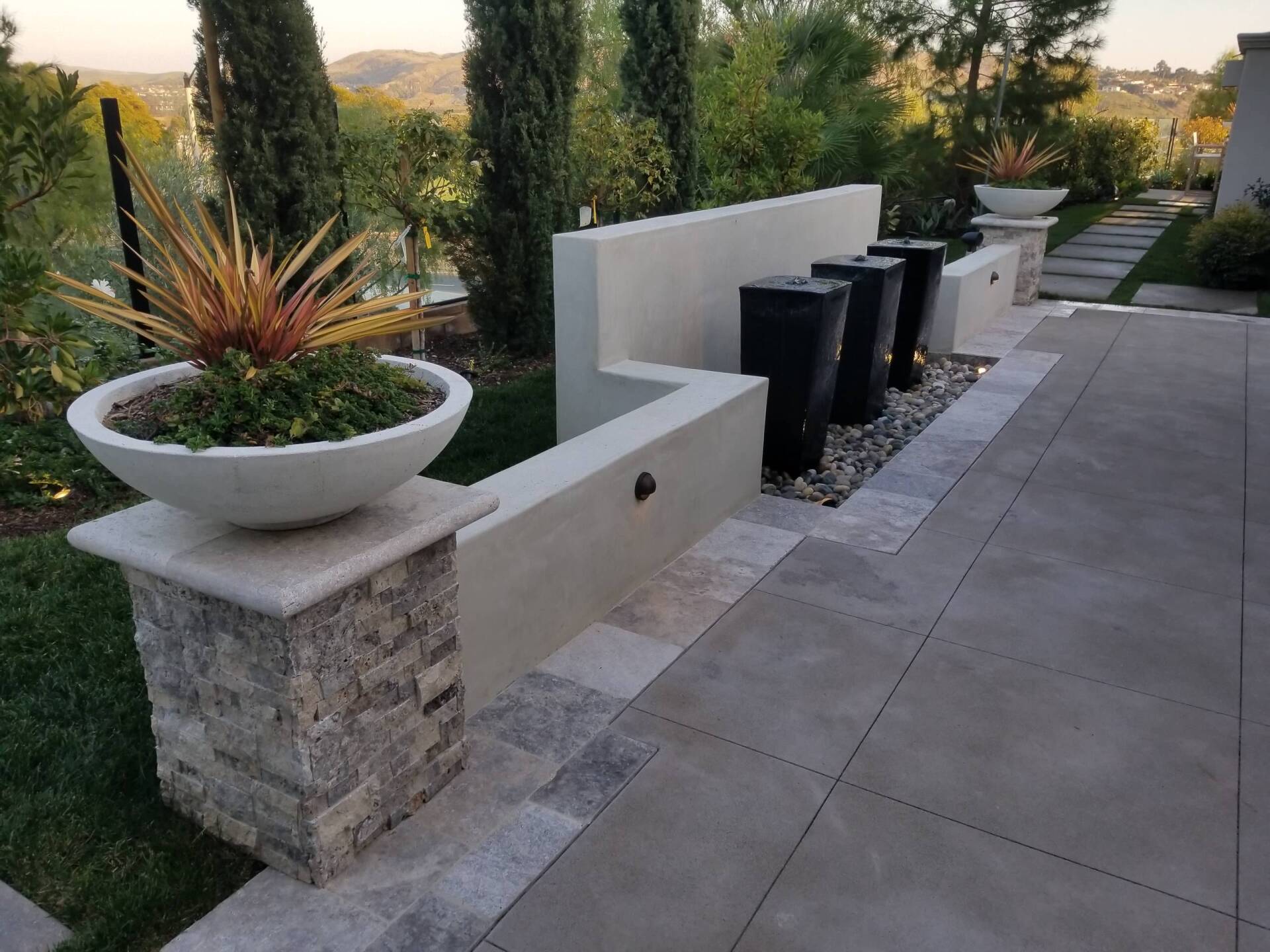 A modern outdoor water feature with planters and stone columns, set against greenery and a clear sky.