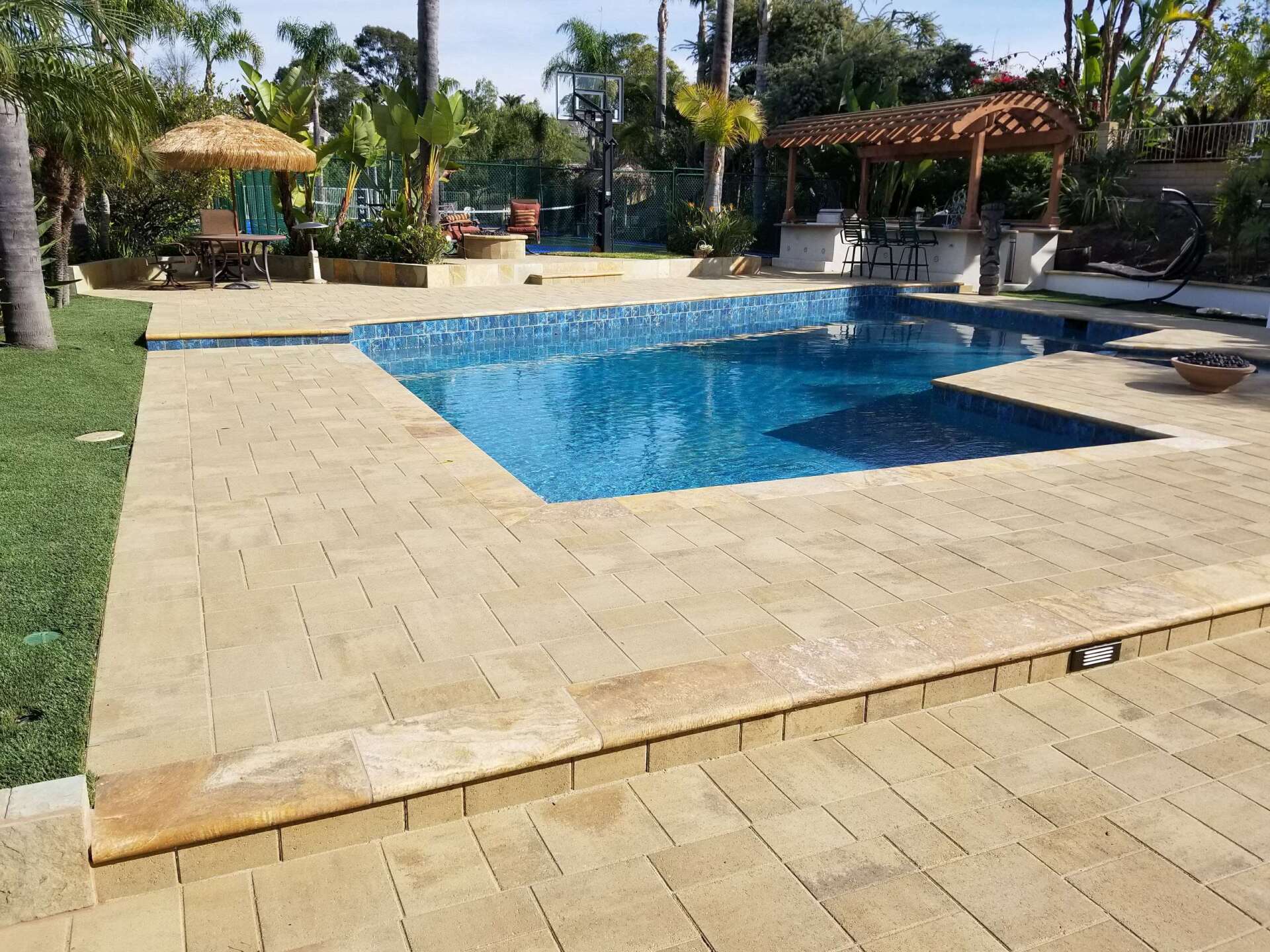 A swimming pool surrounded by tan pavers, with a patio area and lush greenery in the background.