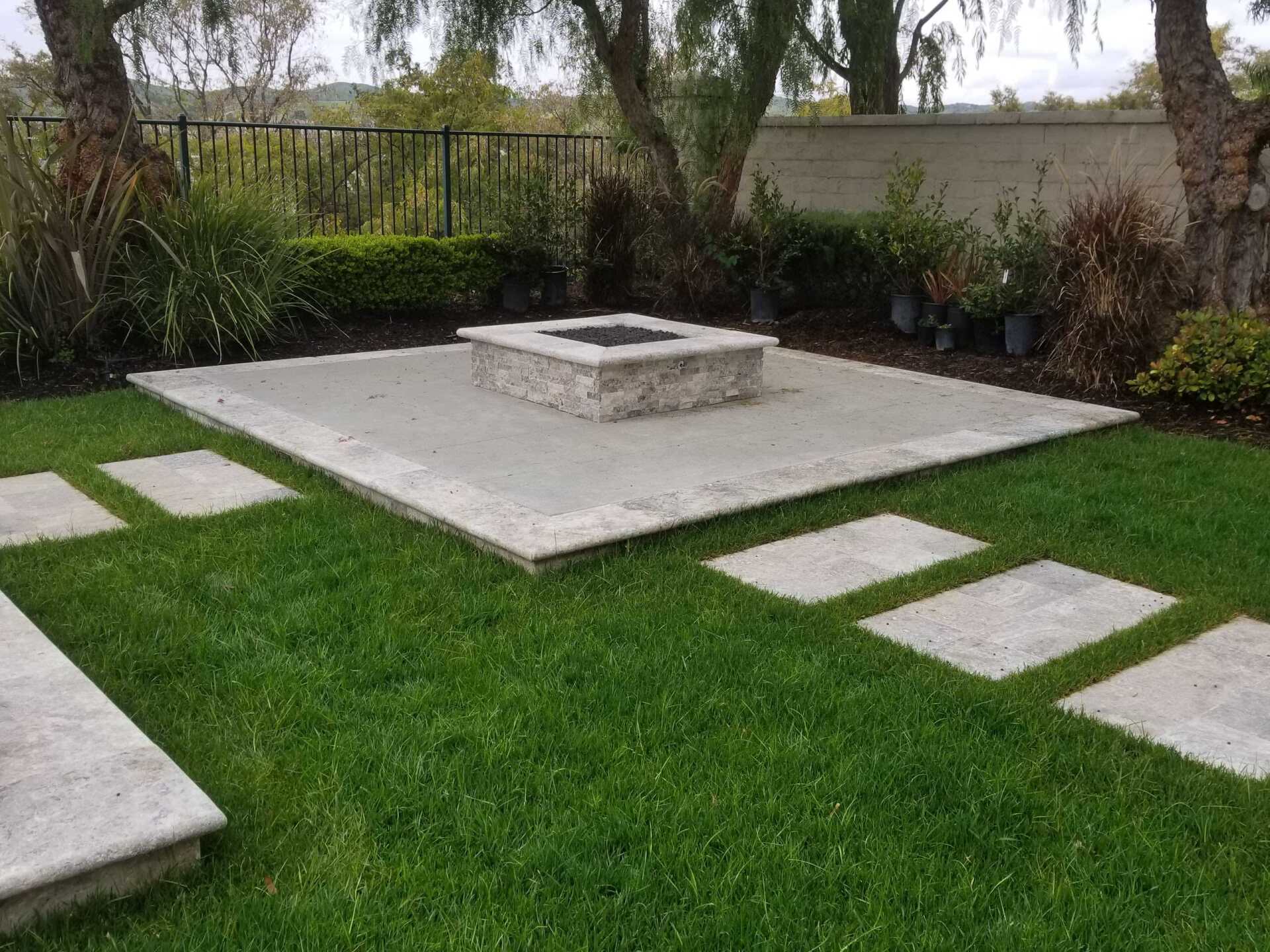 Concrete patio with a fire pit, stepping stones on green grass, surrounded by landscaping.