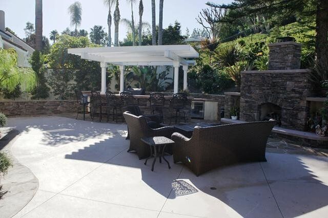 Outdoor patio with a bar, seating area, and fireplace, set in a sunny backyard.
