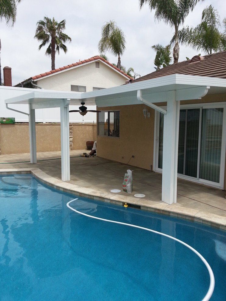 Patio cover over a pool. White columns and roof, blue water, beige house, palm trees.