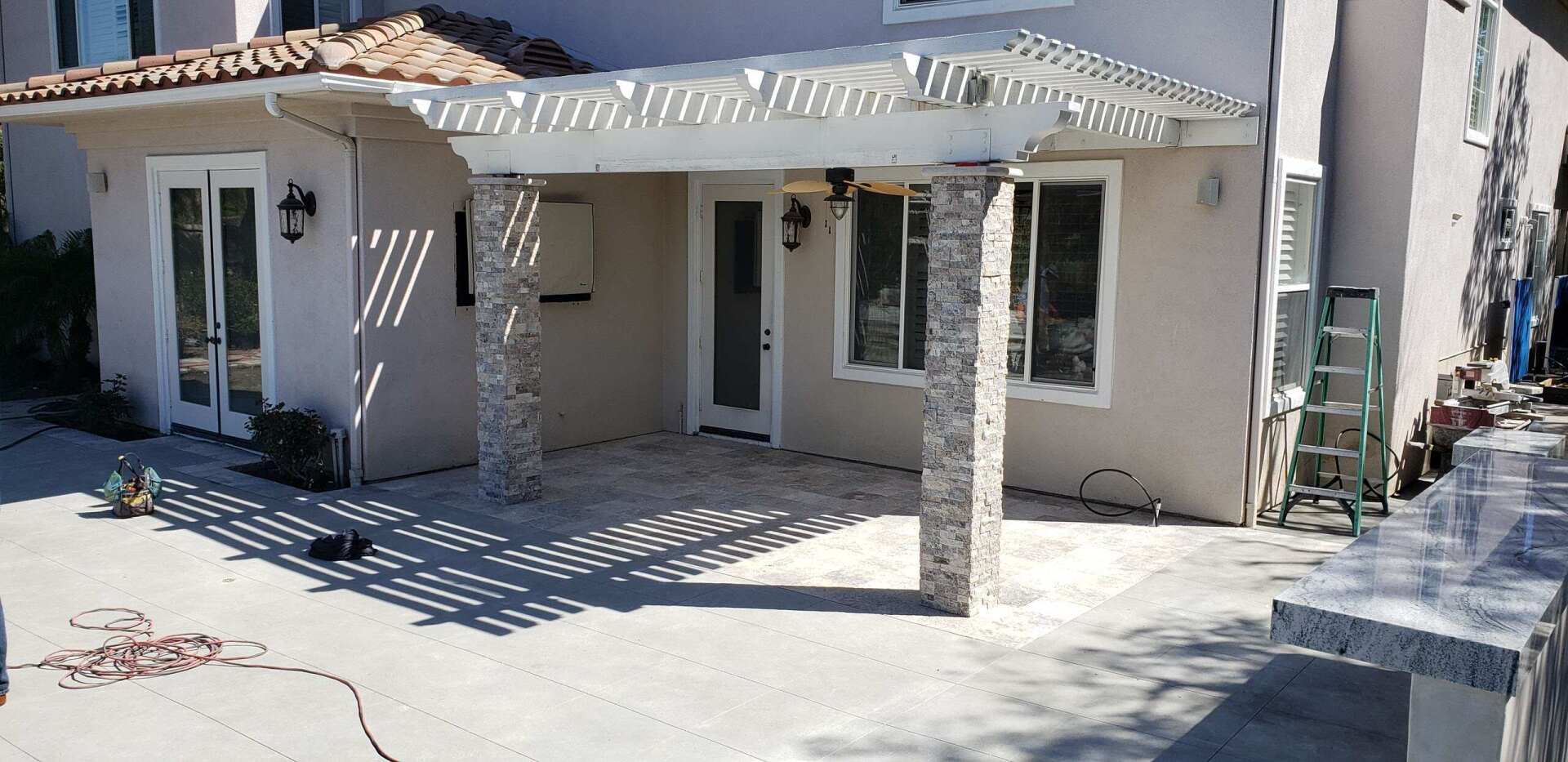 A patio with a pergola. Stone columns and a light-colored house with windows and doors.