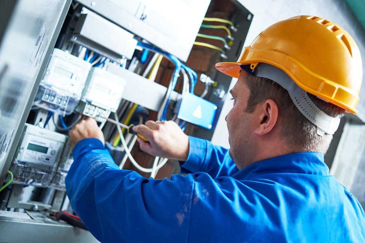 An electrician wearing a yellow hard hat and blue overalls is fixing circuit breakers.