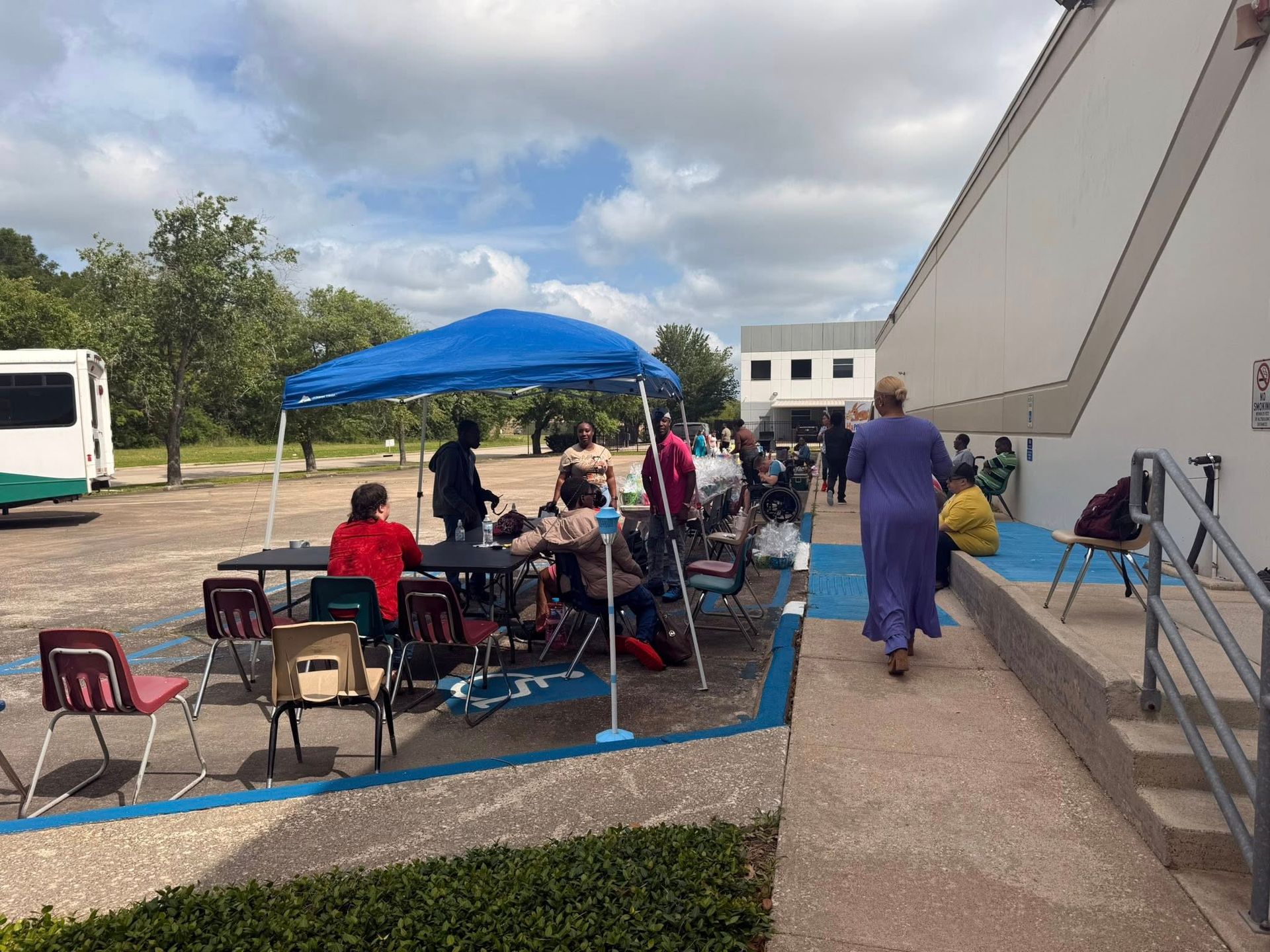 A group of people are sitting under a blue tent in a parking lot.
