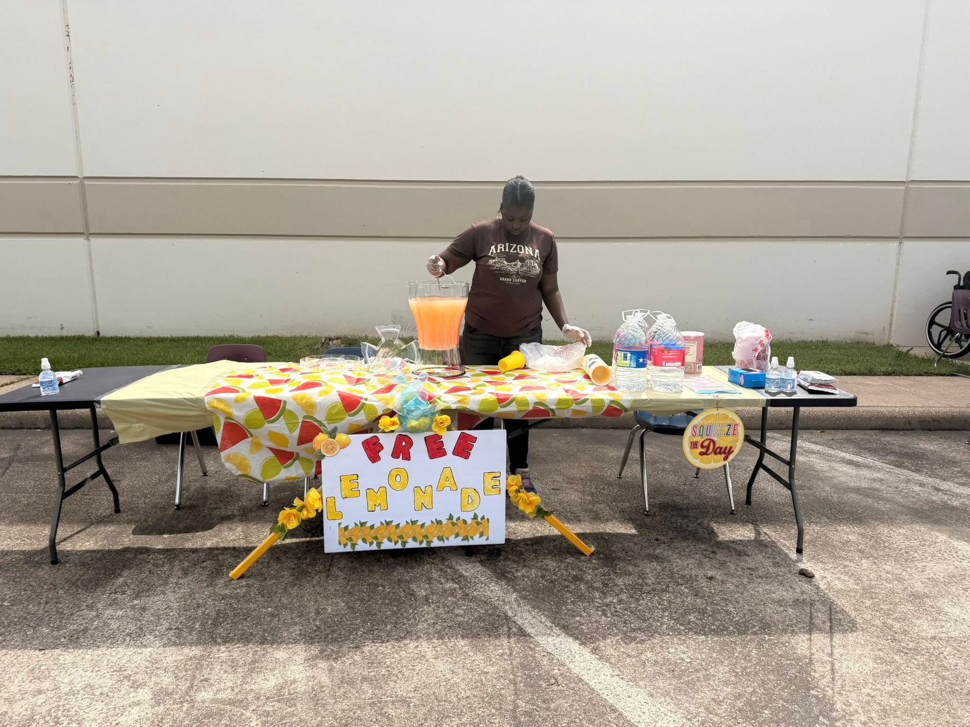 A woman is standing behind a table selling lemonade in a parking lot.