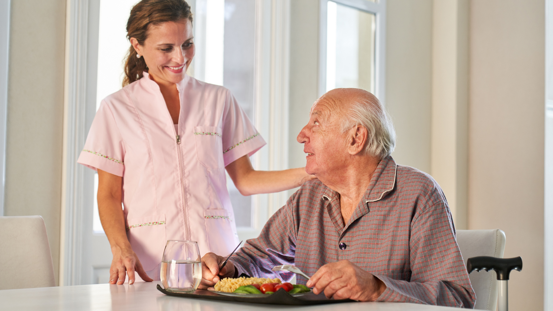 A caregiver in pink scrubs smiles at an elderly man seated at a table, assisting with his meal.