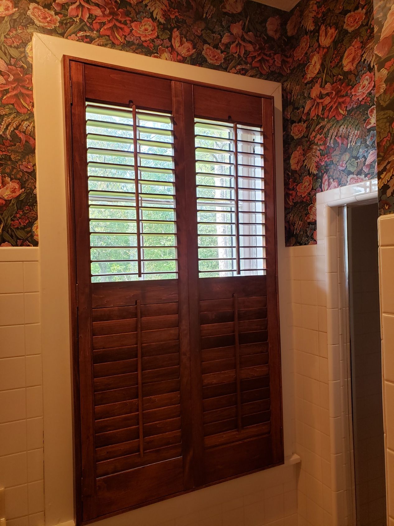 A window with wooden shutters in a bathroom with floral wallpaper.