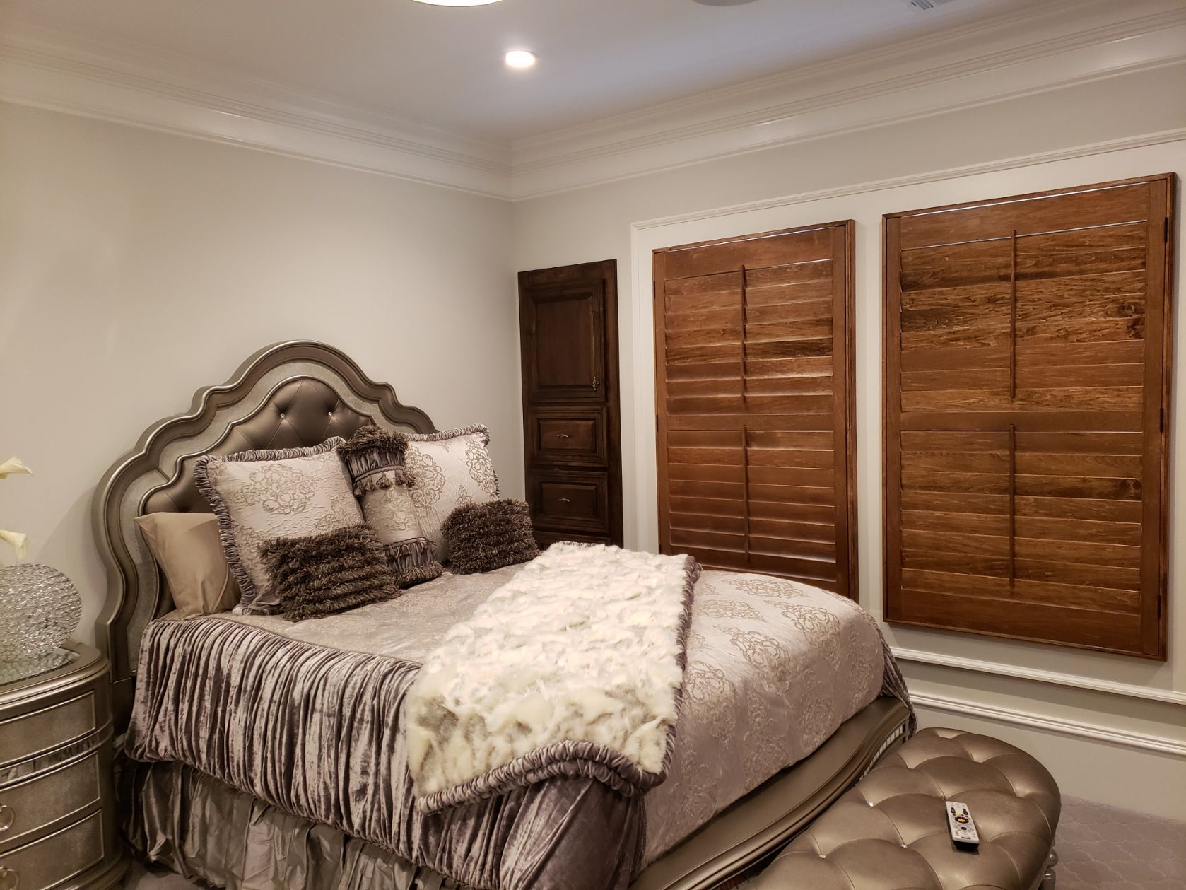 A bedroom with a bed , nightstand , ottoman and wooden shutters.
