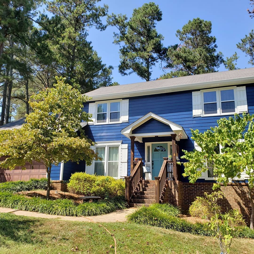 Blue house with white trim, brown steps, and green landscaping. Trees surround the house on a sunny day.