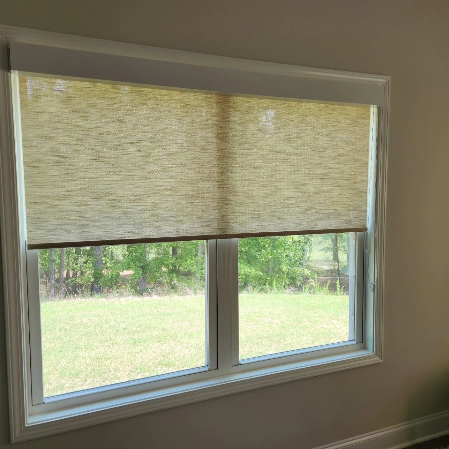 Window with beige roller shade, revealing a grassy yard and trees outside.
