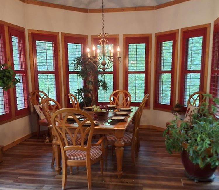 Dining room with wood furniture, bay windows with red frames and shutters, chandelier.