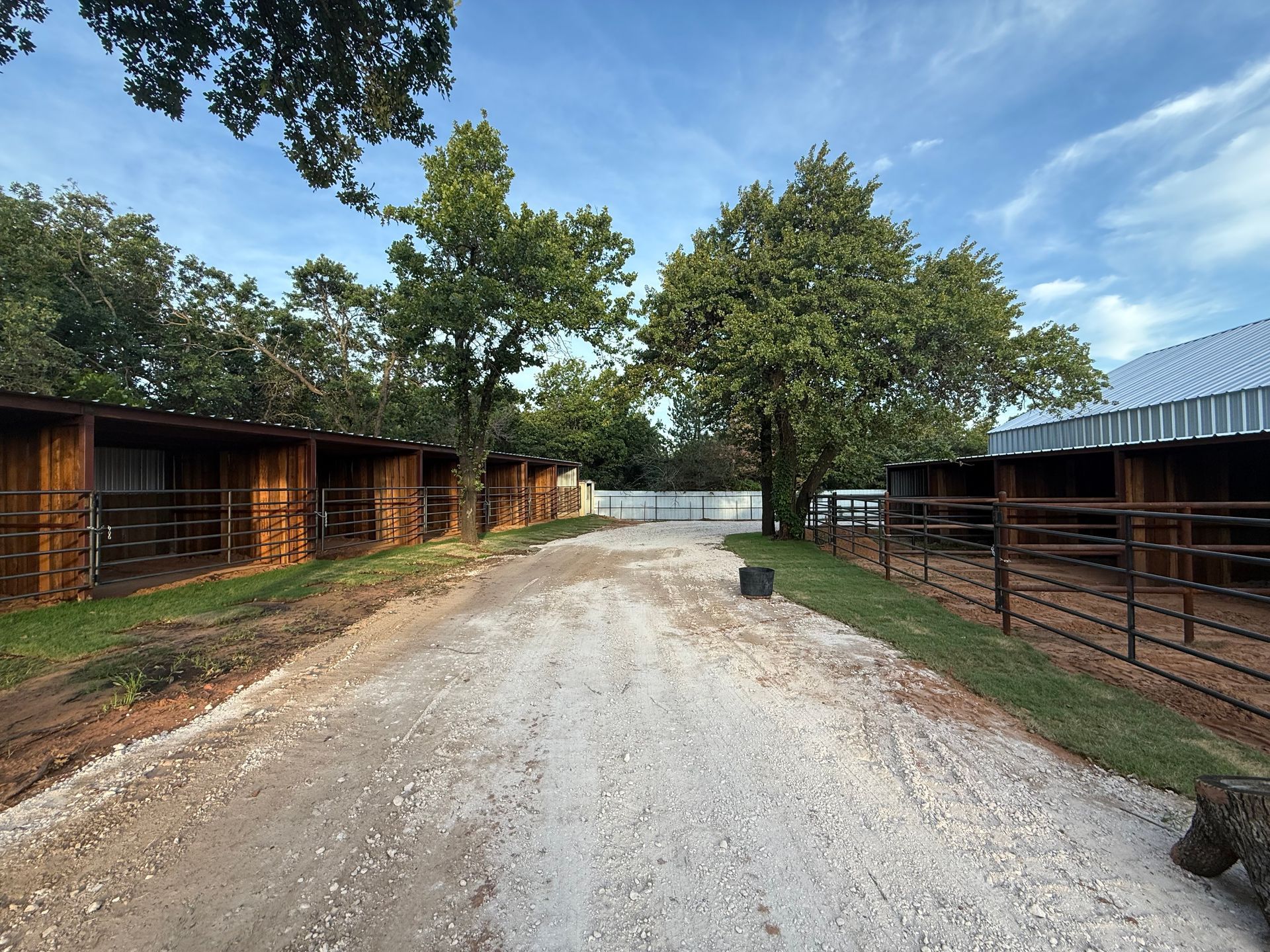 A gravel road between stables and fenced horse stalls under a blue sky.