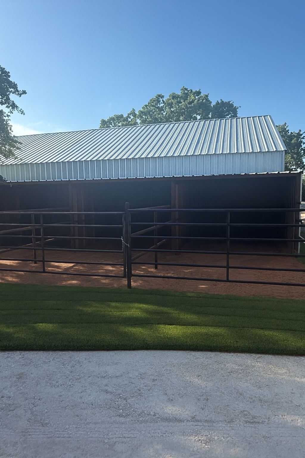 Metal horse stalls with a metal-roofed barn in the background, set against a bright blue sky.
