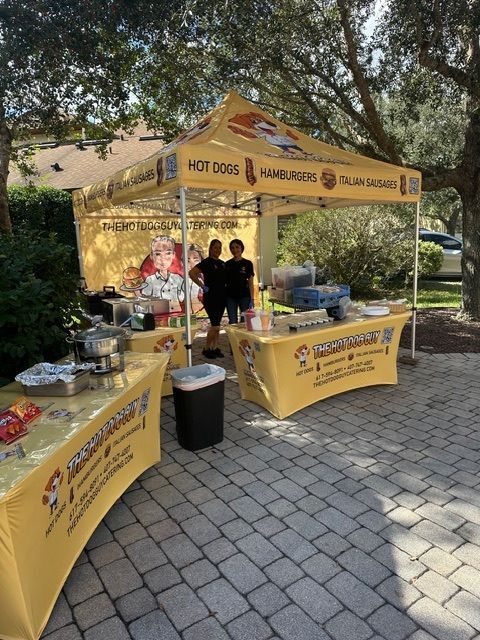 Two people stand behind a yellow, branded pop-up food stand offering hot dogs and hamburgers on a paved patio.