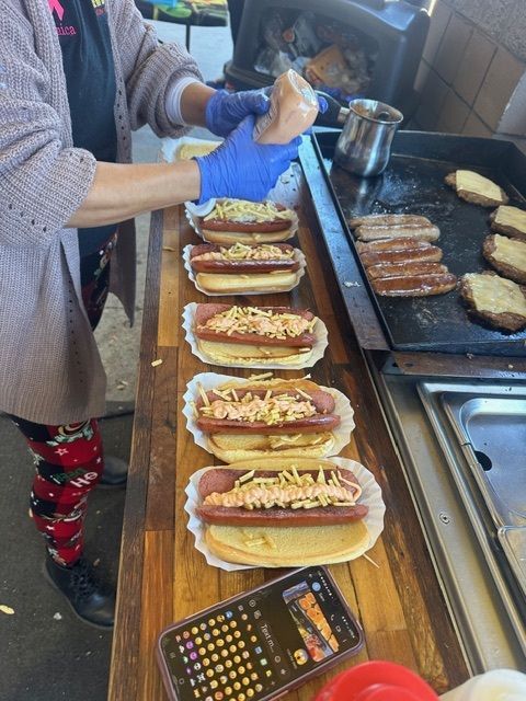 A person wearing blue gloves squirts sauce onto hot dogs lined up on a wooden counter next to a flat-top grill.