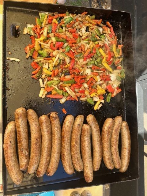 Eleven sausages and a pile of colorful sautéed peppers and onions cooking on a flat-top grill.