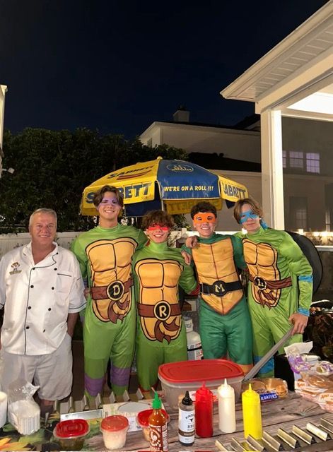 A person in a chef's uniform stands next to four people in Teenage Mutant Ninja Turtle costumes behind a condiment table.