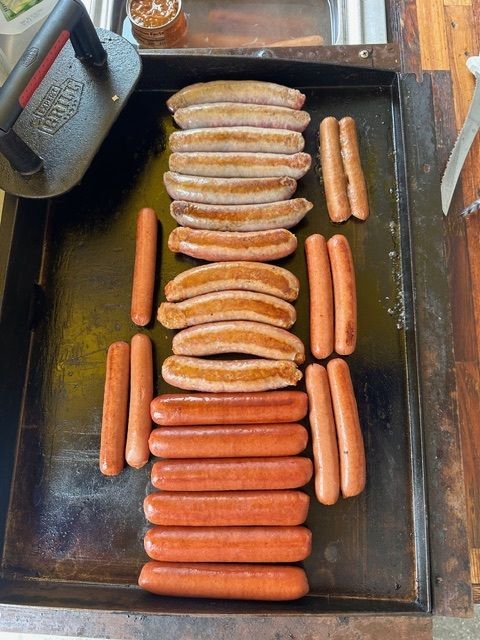 A variety of sausages and hot dogs cooking on a large, oiled flat-top grill.