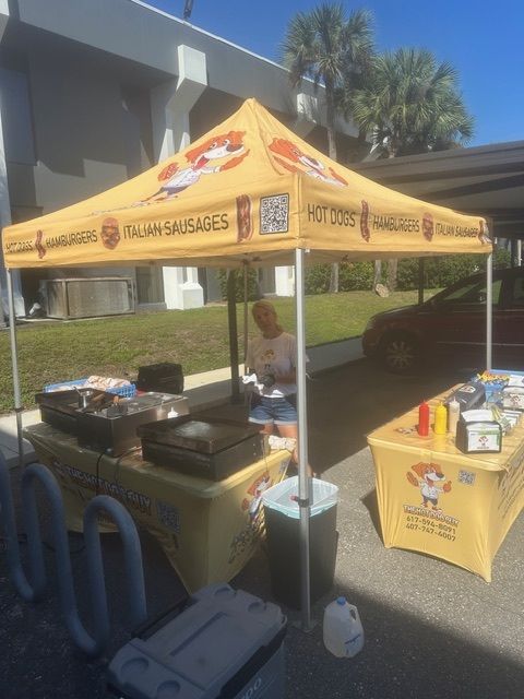 A food vendor stand with a yellow tent and table serving hot dogs and sausages on a sunny outdoor patio.