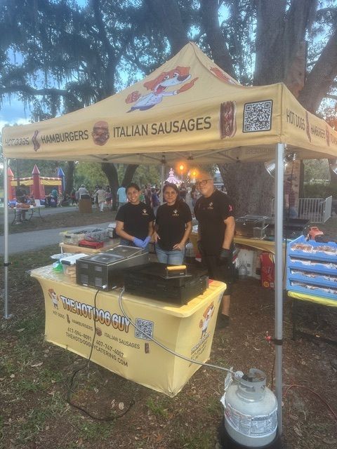 Three people stand behind a yellow food stall tent serving hamburgers and Italian sausages in an outdoor park setting.
