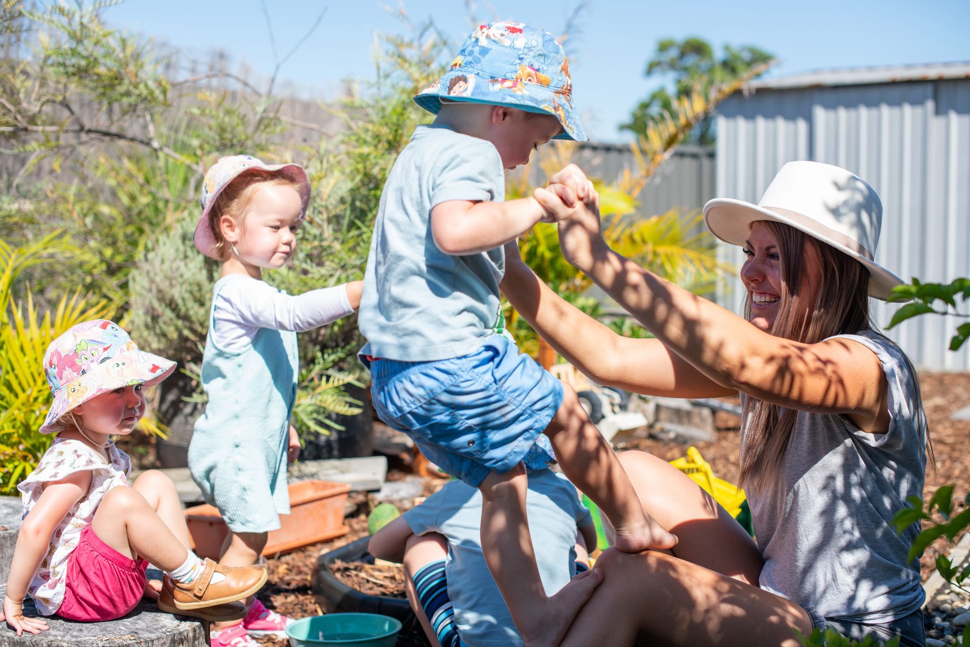 A woman lifts a boy in the air as two other children watch in an outdoor setting, all wearing hats.