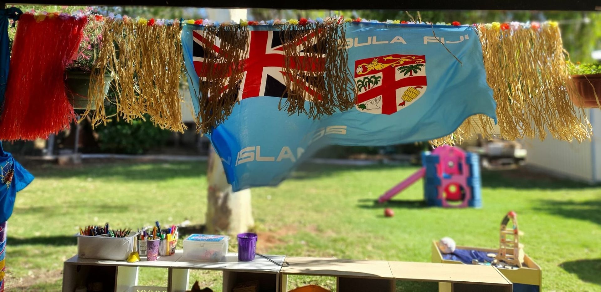 A Fiji flag hanging on a clothesline in a backyard with craft supplies and a playground.