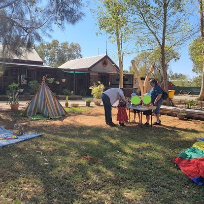 People and children in a grassy yard by a building with a teepee and table.