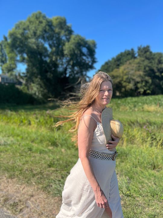 Vrouw in een linnen  jurk staat in een zonnig grasveld met bomen en een blauwe lucht op de achtergrond.