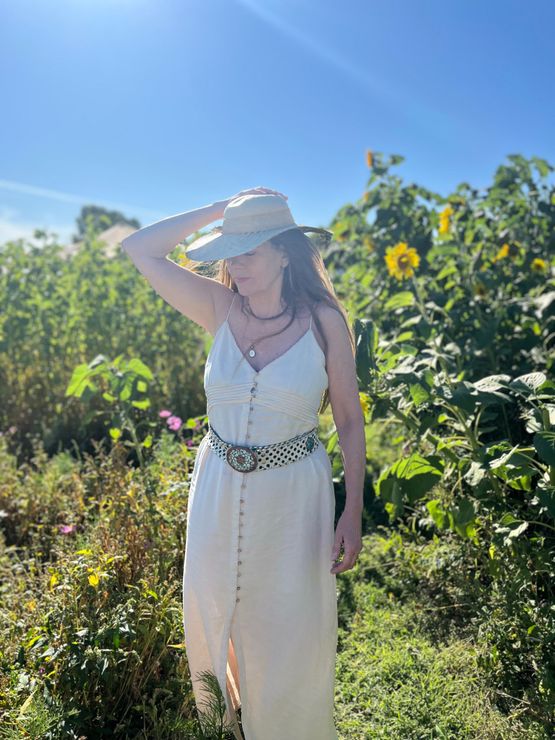 Sieta Malvatherapeut Een vrouw in een witte jurk en een strohoed staat in een zonnige tuin en houdt de rand van haar hoed vast.