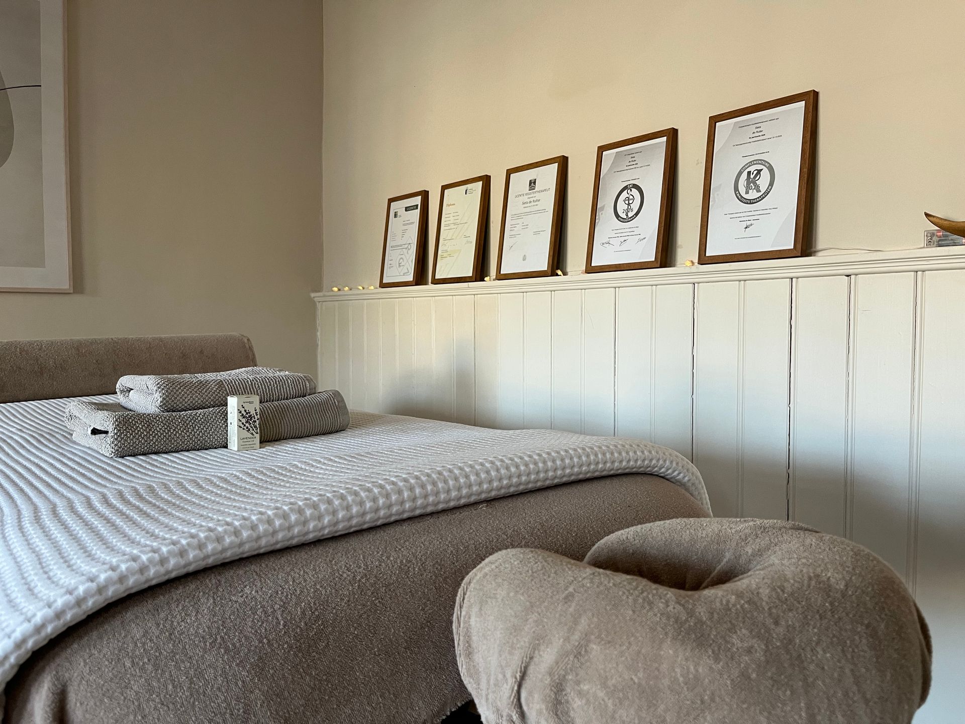 massagetafel Gezellige slaapkamer met een bed, beige sierkussens en ingelijste prenten aan een witte lambrisering.