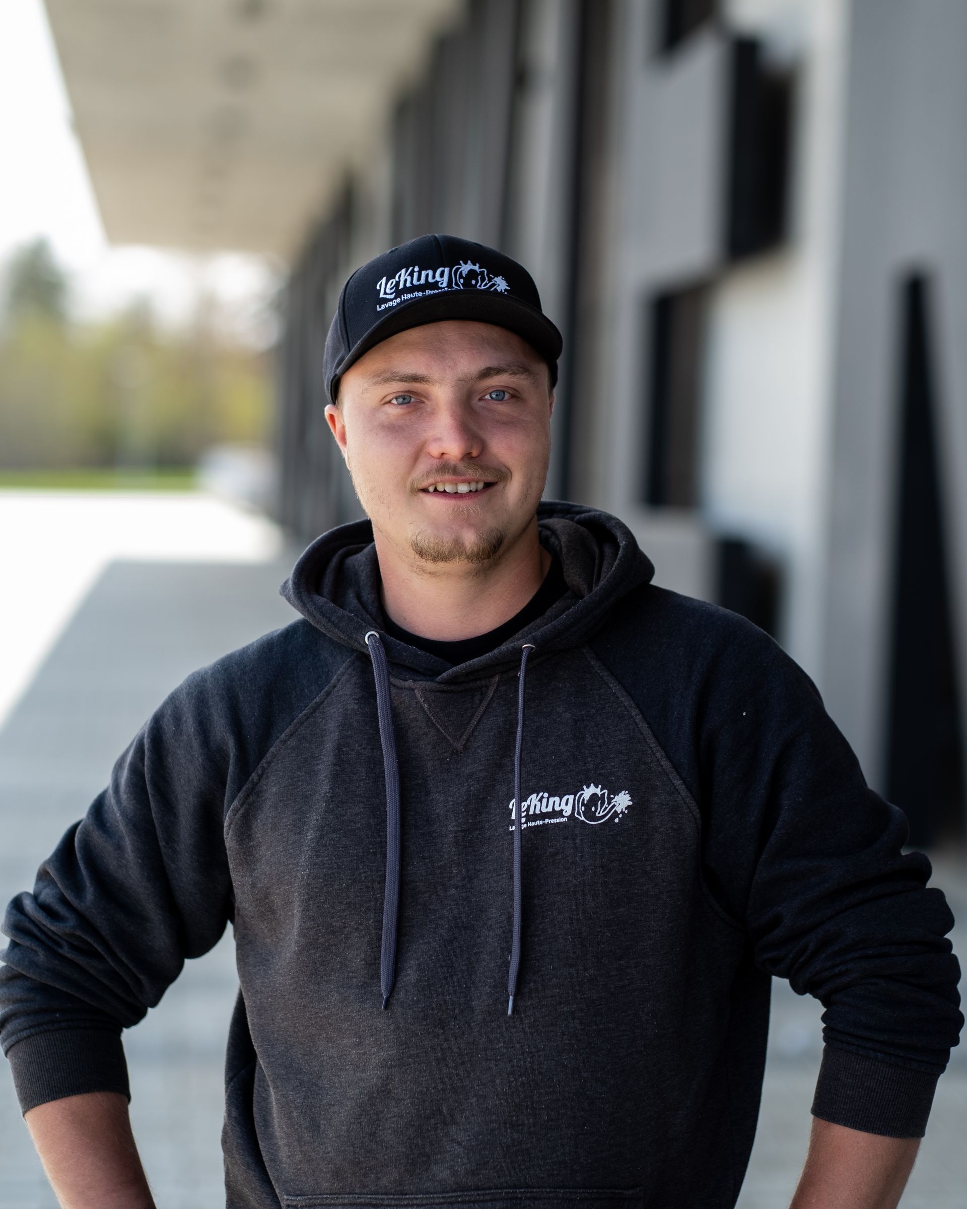 Un homme portant un chapeau et un sweat à capuche se tient debout, les mains sur les hanches.