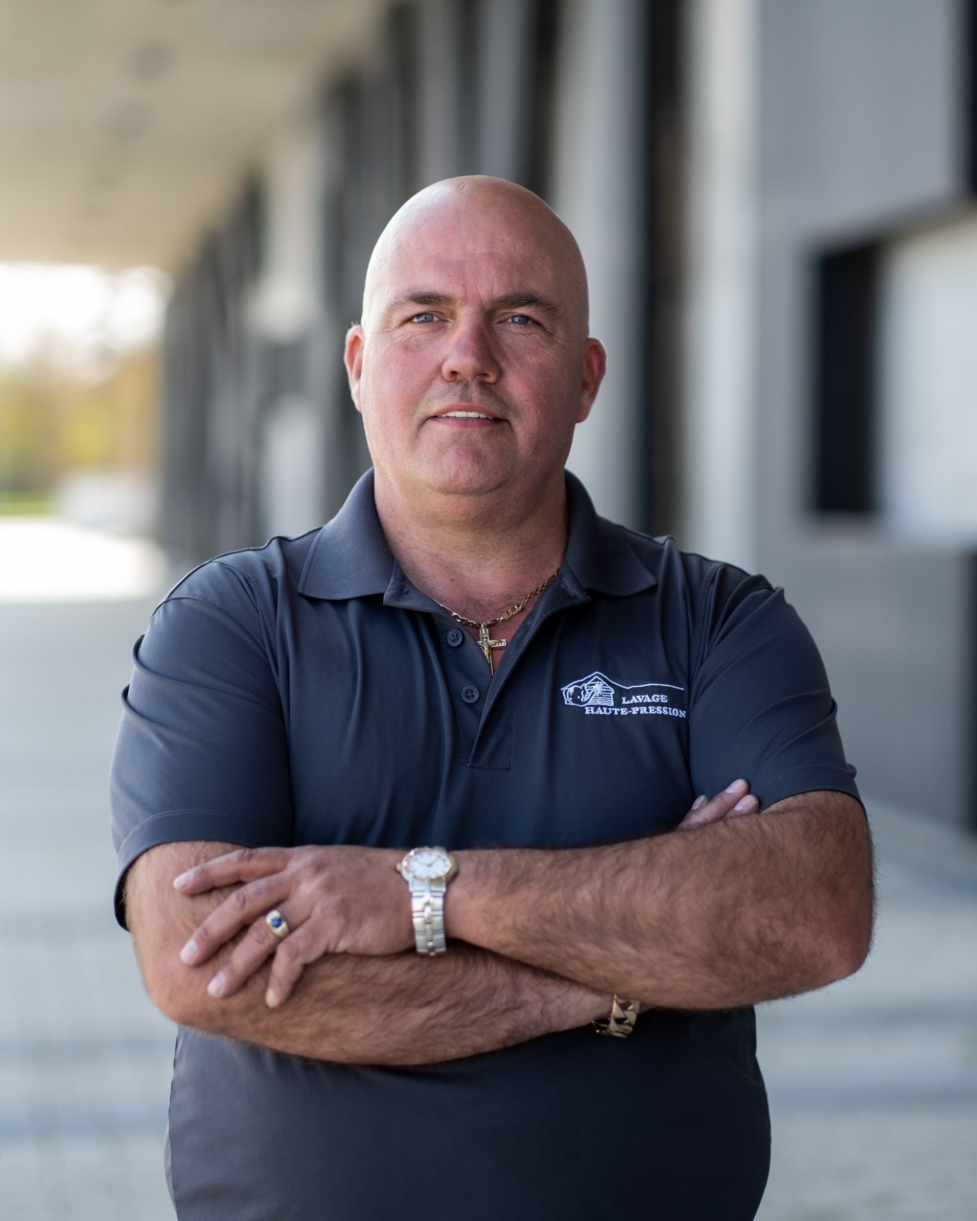 Un homme chauve se tient debout, les bras croisés, devant un immeuble.