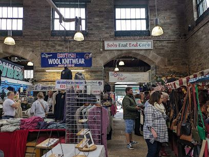 Market interior with people browsing stalls selling wool knitwear, bags, and food.