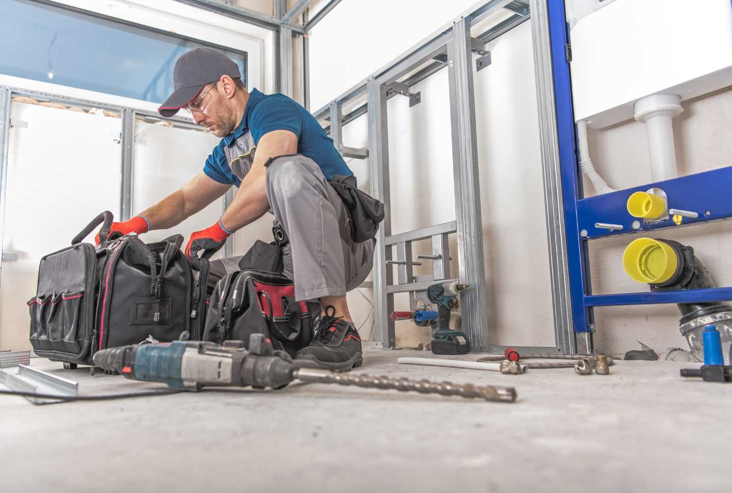A man is kneeling down in a bathroom with a tool bag and a drill.