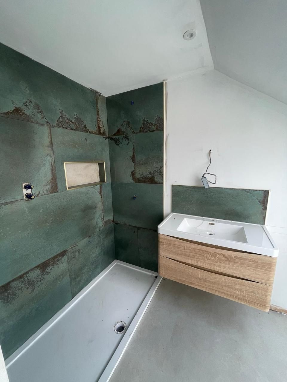 A bathroom under construction featuring dark green stone-look wall tiles, a white shower tray, and a wall-mounted wood vanity.