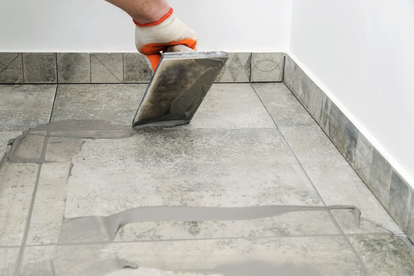 Person using a trowel to apply grout between floor tiles, wearing an orange glove.