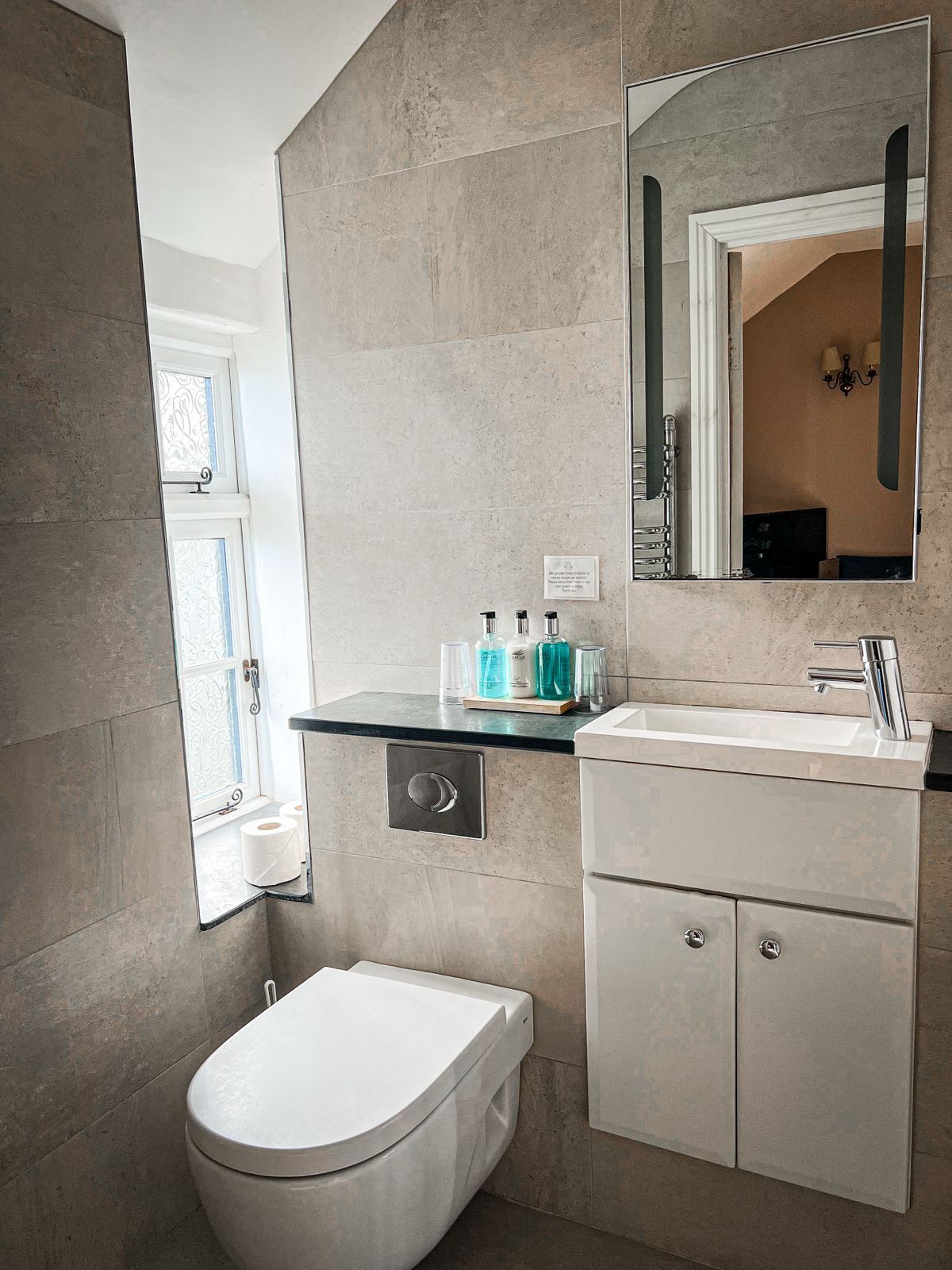 A small modern bathroom with beige stone-tiled walls, a wall-mounted toilet, vanity sink, and a framed mirror.