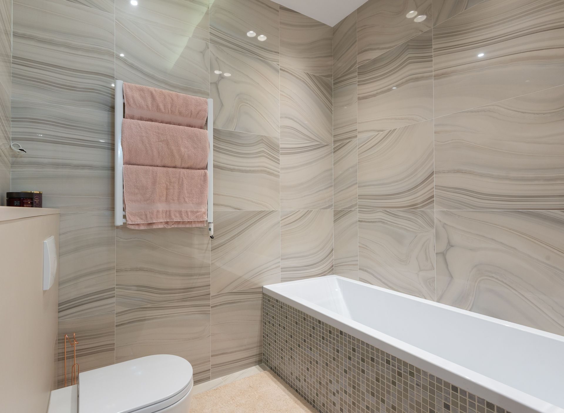 Modern bathroom with marble-patterned walls, white bathtub, towel rack, and toilet underneath.