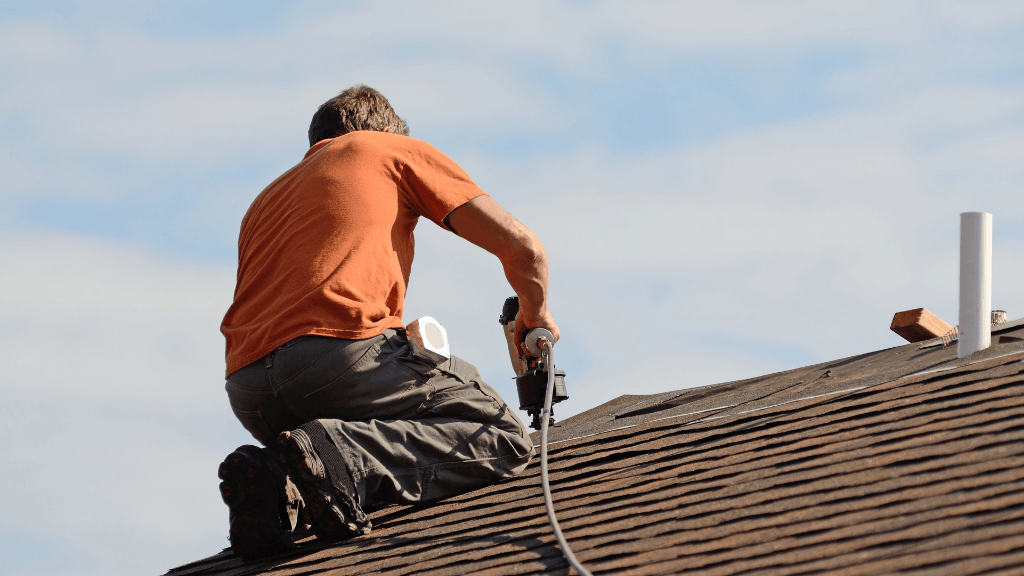 A man is kneeling on a roof using a drill.