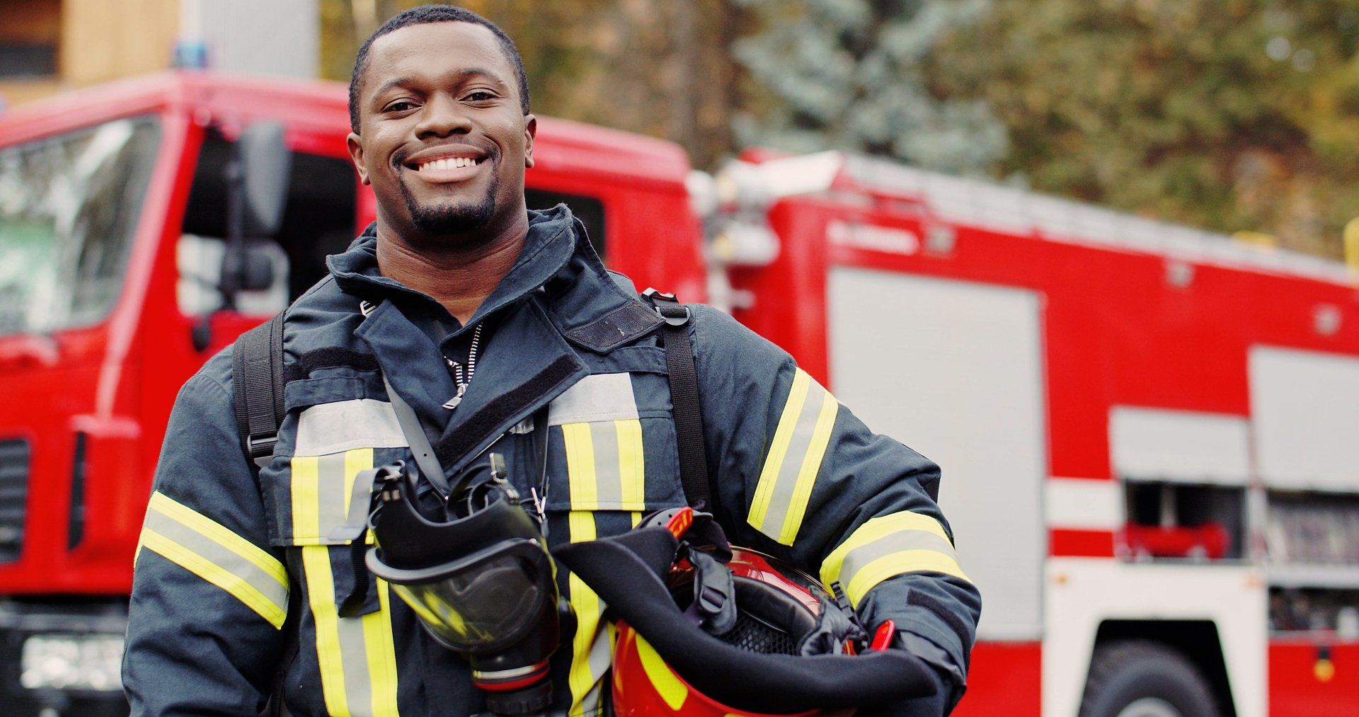 A fireman is standing in front of a fire truck holding a helmet and smiling.