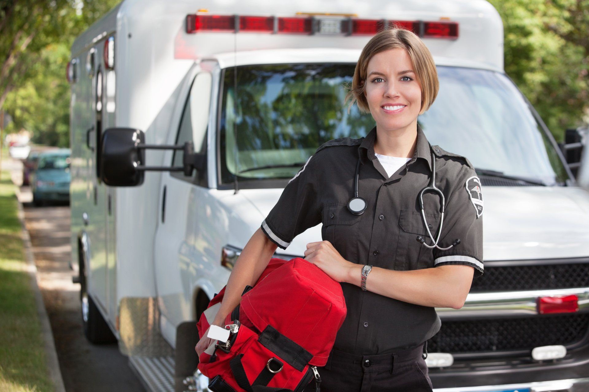 A female paramedic is standing in front of an ambulance holding a bag.