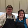 A man and a woman wearing aprons are posing for a picture in a kitchen.