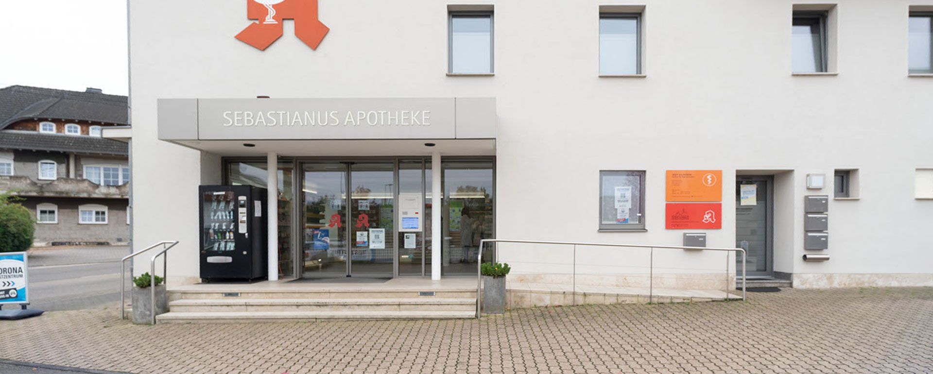 Ein weißes Gebäude mit einem roten Kreuz an der Seite und einem Verkaufsautomaten davor.